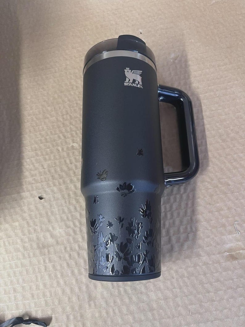 Op de afbeelding: Een donkerblauwe Stanley reismok met een handvat en een bloemenontwerp ge&euml;tst op de bodem. Het Stanley-logo is zichtbaar. De mok heeft een zwart deksel en is ontworpen om dranken warm of koud te houden.
