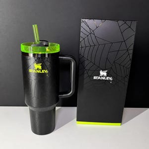 May include: Black Stanley tumbler with a neon green lid and straw, and a matching box. The tumbler features a spiderweb design and the Stanley logo in yellow. The box also has a spiderweb design.