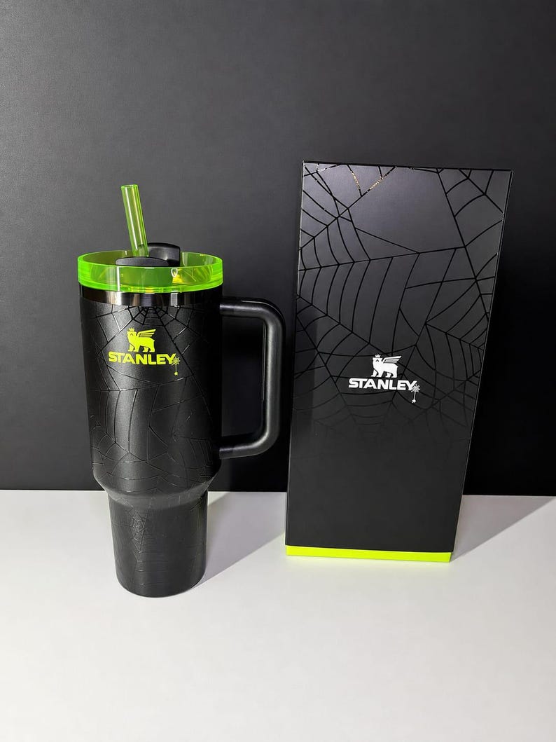 May include: Black Stanley tumbler with a neon green lid and straw, and a matching box. The tumbler features a spiderweb design and the Stanley logo in yellow. The box also has a spiderweb design.