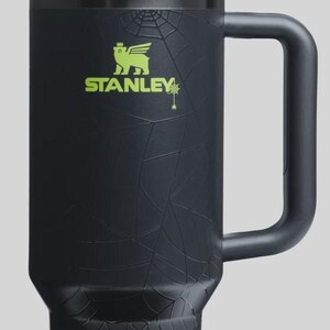 May include: Black Stanley tumbler with a green lid and straw. The tumbler features a spiderweb design and the Stanley logo in green. The handle is black. A Halloween-themed drinkware item.