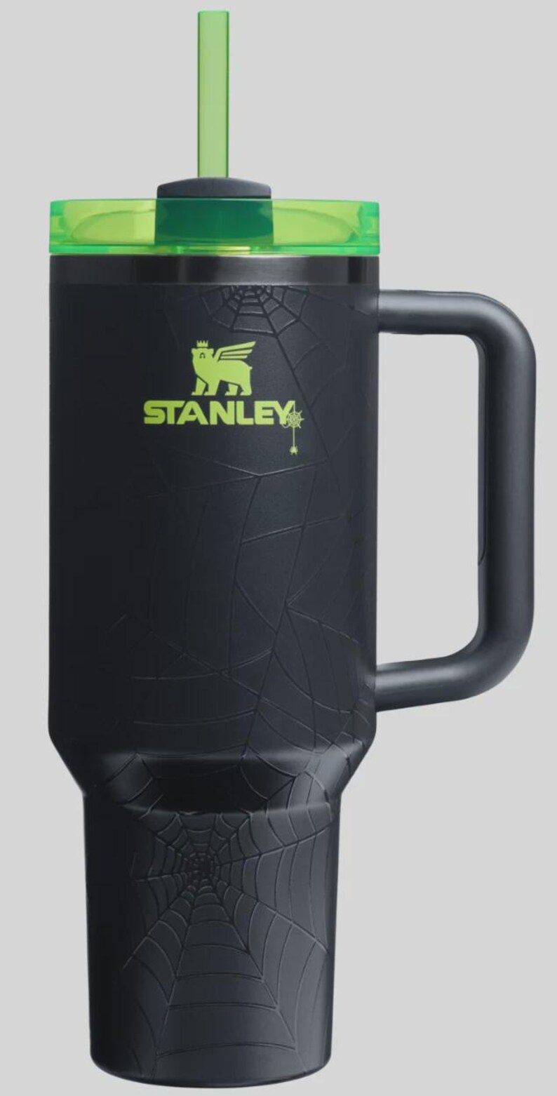 May include: Black Stanley tumbler with a green lid and straw. The tumbler features a spiderweb design and the Stanley logo in green. The handle is black. A Halloween-themed drinkware item.