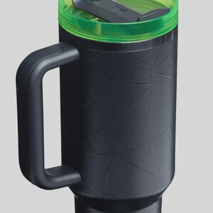 May include: Black travel mug with a green lid and straw. The mug has a handle and a textured spiderweb design. The lid is translucent green, and the straw is a matching green color. The mug is designed to keep drinks cold or hot.