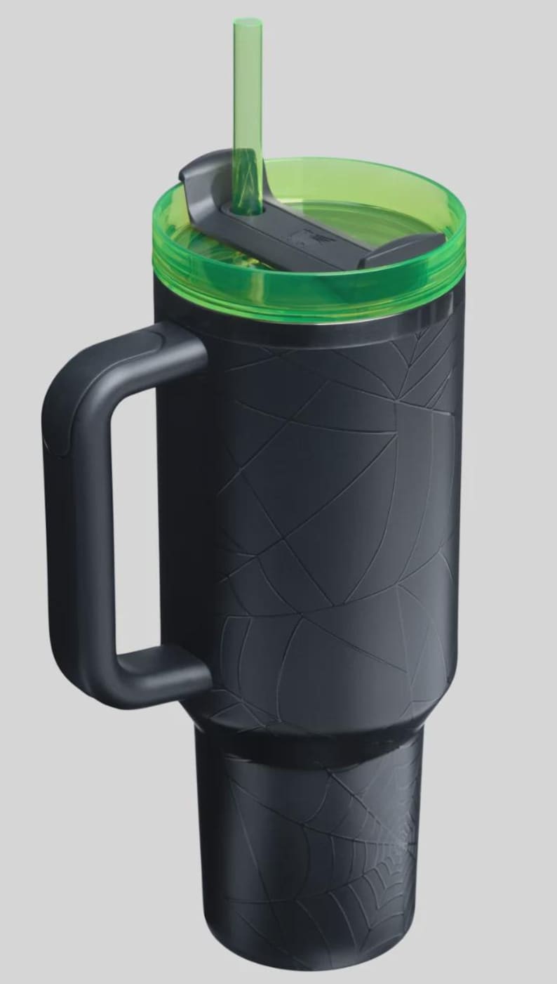 May include: Black travel mug with a green lid and straw. The mug has a handle and a textured spiderweb design. The lid is translucent green, and the straw is a matching green color. The mug is designed to keep drinks cold or hot.