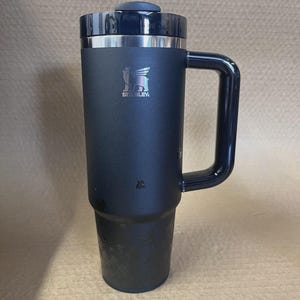 Op de afbeelding: Een zwarte ge&iuml;soleerde reismok met een handvat en een deksel. De mok heeft een zilveren rand en het Stanley-logo. De mok is ontworpen om dranken langdurig warm of koud te houden.
