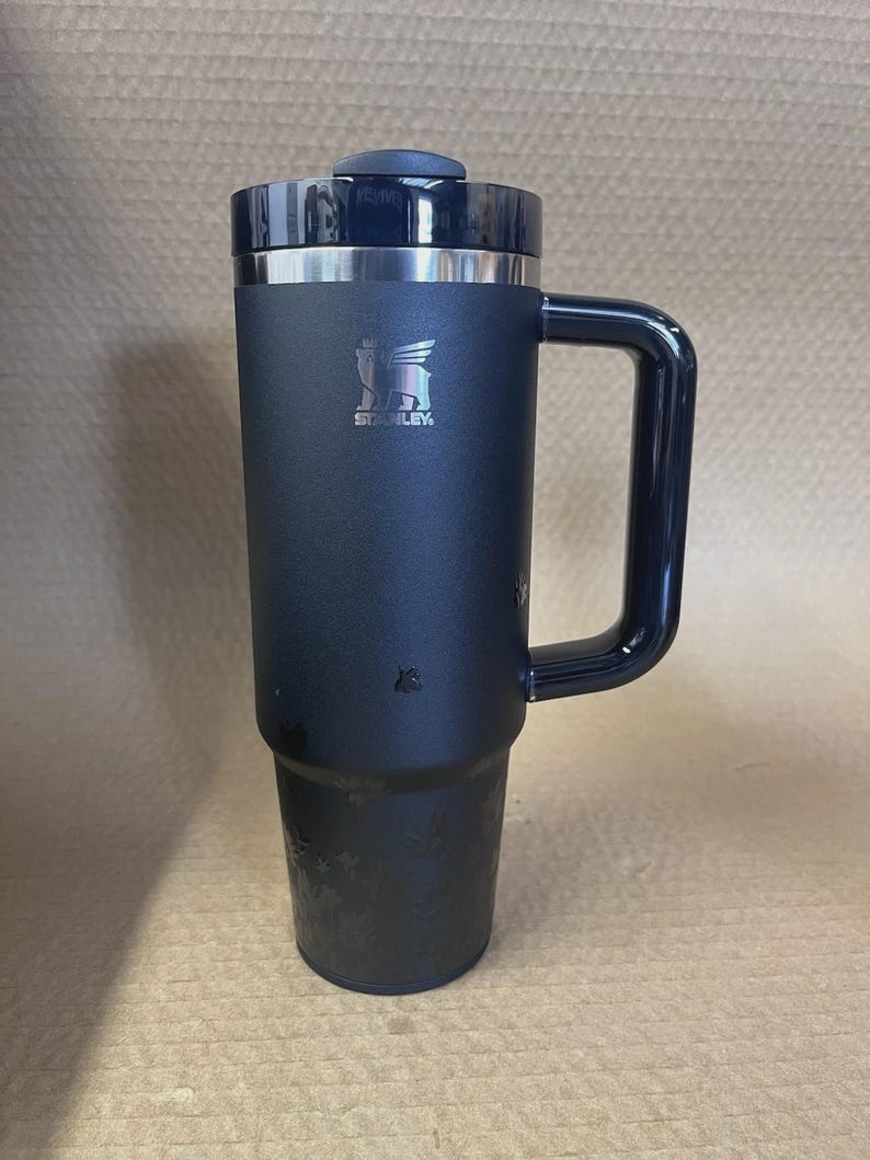 Op de afbeelding: Een zwarte ge&iuml;soleerde reismok met een handvat en een deksel. De mok heeft een zilveren rand en het Stanley-logo. De mok is ontworpen om dranken langdurig warm of koud te houden.