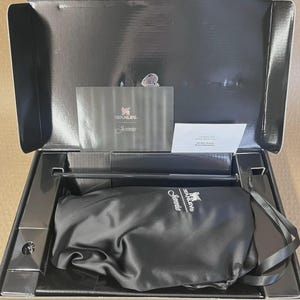 Op de afbeelding: Een open zwarte doos met een Stanley-product. Binnenin is een zwarte satijnen zak met het Stanley-logo en de naam "Jamie" zichtbaar, samen met een boekje en een witte kaart. De doos is open en onthult de inhoud.