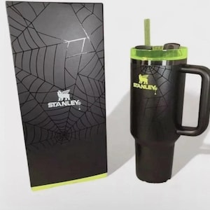 May include: Black Stanley travel mug with a green lid and handle, next to a matching box. The mug and box feature a spiderweb design and the Stanley logo. The mug includes a straw.