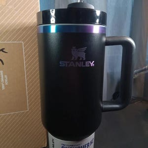 Op de afbeelding: Een zwarte Stanley Quencher H2.0 Flowstate tumbler met een handvat en een rietje. De tumbler heeft een zwart deksel met een kleurrijke iriserende band. De tumbler is 1,18L.