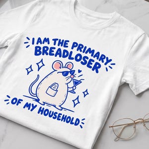 May include: A white short-sleeved t-shirt featuring a blue graphic of a mouse wearing sunglasses, holding a martini glass, and carrying a baguette in a pocket. The text reads I AM THE PRIMARY BREADLOSER OF MY HOUSEHOLD in blue capital letters.