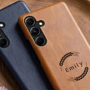 Puede incluir: Dos fundas de teléfono, una azul marino y otra marrón claro, sobre una bandeja de madera. La funda marrón claro tiene el nombre "Emily" impreso, rodeado de un diseño de trigo. Pétalos de flores rosas esparcidos.
