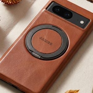 以下が含まれることがあります： 黒いリングホルダーが付いた茶色の革製携帯電話ケース。「JAMES」の文字がリングホルダーにプリントされています。携帯電話ケースにはカメラの切り抜きがあり、ドライフラワーに囲まれています。