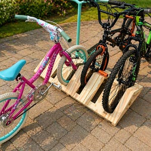 May include: A wooden bike rack holds three bicycles. A pink child's bike with a blue seat and white tyres is in the front. Two black mountain bikes with orange and green accents are behind it. The rack is on a brick patio.