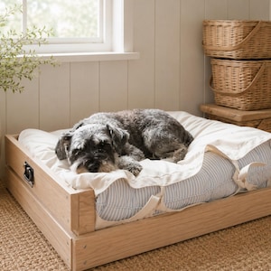 May include: A dog bed with a light wood frame, featuring a white and blue striped pillow and a white blanket. A small dog with gray and black fur is resting on the bed. Wicker baskets are stacked in the background.