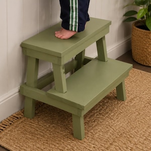 May include: A sage green wooden step stool with two steps. The stool is placed on a woven jute rug. A person's foot is on the top step. A potted plant in a woven basket is in the background.