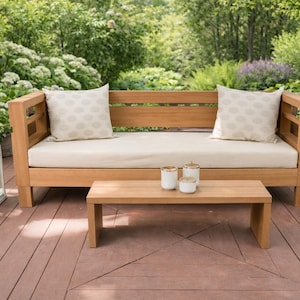 Op de afbeelding: Een houten tuinmeubelset met een bank met twee kussens met patronen, een lage salontafel en twee witte lantaarns. De bank en tafel zijn gemaakt van lichtgekleurd hout, geplaatst op een houten terras, met groen op de achtergrond.
