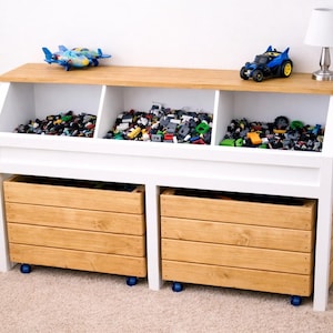 May include: A white and wood toy storage unit with three open compartments on top filled with toys. Two wooden drawers are below, and a blue toy airplane, a blue toy car, and a small lamp sit on top.