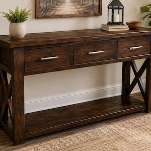 Puede incluir: Una mesa consola de madera marrón oscuro con tres cajones y tiradores horizontales de metal plateado. La mesa cuenta con una superficie rectangular, un estante inferior y soportes laterales en forma de X con veta de madera visible.