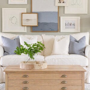 May include: A living room scene featuring a white sofa with blue pillows, a wooden coffee table, and framed artwork on the wall. The coffee table has three drawers and a round wooden tray. Green plants in a glass vase add a natural touch.