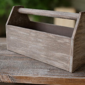May include: A rustic wooden toolbox with a handle. The box is painted in a distressed gray finish, giving it a weathered appearance. The toolbox is empty and sits on a wooden surface.