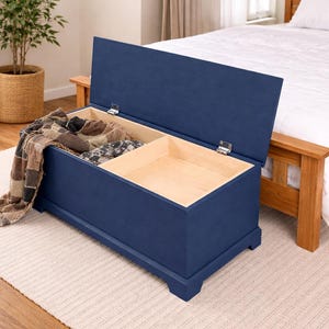 May include: A navy blue storage chest with a hinged lid, partially open to reveal a light wood interior. A patterned blanket and other items are inside. The chest is on a light-colored rug near a bed.