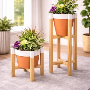 Puede incluir: Dos soportes de plantas de madera con macetas naranjas y blancas llenas de vegetación y flores moradas. Los soportes tienen un acabado de madera natural y están colocados sobre una alfombra gris. Un soporte es más alto que el otro.