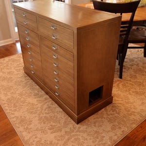 May include: A large, wooden cabinet with multiple drawers and a rectangular shape. The cabinet has a light brown finish and silver drawer pulls. It is placed on a patterned rug in a room with a wooden floor.