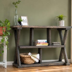 May include: A dark wood console table with an X-frame design, featuring two shelves. The top shelf holds a glass vase, a small potted plant, and a framed sign that says hi. The middle shelf contains a stack of books and a small box. A woven basket sits on the bottom shelf.