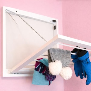 May include: A white, wall-mounted drying rack with items hanging on it. The rack is open, displaying a gray scarf with white pom-poms, a blue towel, and blue gloves. The background is a pink wall.