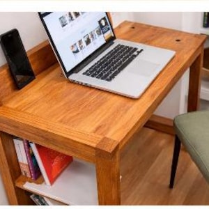 May include: A wooden desk with a laptop, smartphone, and books. The desk is made of light brown wood and has a built-in shelf. A laptop is open on the desk, displaying a website. A black smartphone is propped up on the desk.