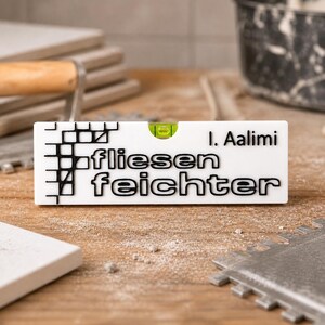 Könnte beinhalten: Ein weißes Wasserwaage mit schwarzem Text "fliesen feichter" und "I. Aalimi". Die Wasserwaage hat eine grüne Libelle. Das Bild zeigt auch Werkzeuge und Materialien zum Fliesenlegen.