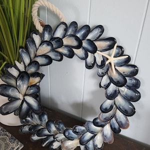 Op de afbeelding: Een cirkelvormige krans gemaakt van blauwe en witte mosselschelpen, met een natuurlijk touw om op te hangen. Een kleine witte zeester is bevestigd. Decoratie voor een maritieme sfeer.