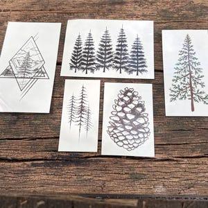 May include: A collection of five temporary tattoos featuring black ink illustrations. The designs include a pine tree inside a geometric diamond shape, a row of five pine trees, two thin pine trees, a detailed pine cone, and a single tall pine tree.