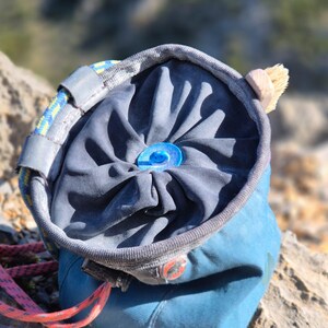 May include: A blue chalk bag for rock climbing, with a drawstring closure and a small brush attached. The bag is made of durable fabric and features a blue plastic closure. A climbing rope is visible.