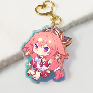 Può includere: Un portachiavi colorato in stile anime con un personaggio dai capelli rosa e occhi viola. Il personaggio indossa un abito rosso e bianco con dettagli dorati. Il portachiavi ha una chiusura a forma di cuore color oro.