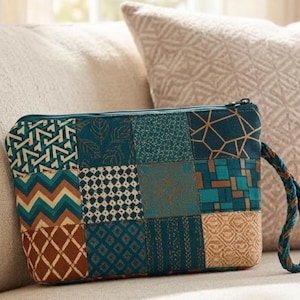 May include: A rectangular zippered pouch featuring a patchwork design of various teal, brown, and tan fabric squares with geometric, leaf, and chevron patterns. The pouch includes a braided teal and brown wrist strap attached to the side zipper.