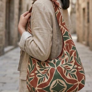 May include: A hobo-style shoulder bag featuring a repeating geometric and botanical pattern in shades of terracotta red, forest green, and cream. The bag has a wide, integrated strap that is tied in a knot at the top, resting on the shoulder.