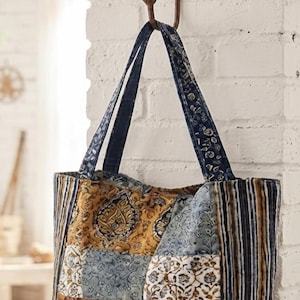 May include: A quilted tote bag featuring a patchwork design with various blue, gold, and white floral and geometric patterns. The bag has two matching fabric shoulder straps and is constructed from multiple rectangular panels of printed cotton fabric.