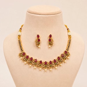 May include: A gold necklace and earring set on a mannequin. The necklace has red, teardrop-shaped stones, green accents, and small white pearls. The earrings match the necklace, with red stones and pearls. The set is designed with a floral pattern.