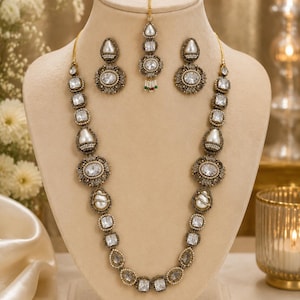 May include: A jewelry set featuring a long necklace, two round earrings, and a maang tikka. The pieces are crafted with antique gold-toned metal, clear rectangular and oval crystals, and white faux pearls, arranged in a symmetrical pattern.