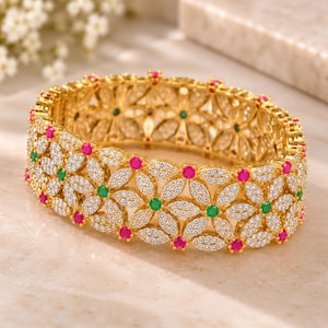 Puede incluir: Un brazalete rígido de tono dorado con un patrón floral repetitivo. Los pétalos están incrustados con pequeñas piedras transparentes, mientras que los centros alternan entre piedras redondas de color rosa y verde. La estructura es calada.