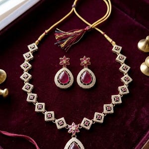 Könnte beinhalten: Ein goldfarbenes Halsketten- und Ohrring-Set mit rubinroten Edelsteinen. Die Halskette hat ein rautenförmiges Design mit einem tropfenförmigen Anhänger. Die Ohrringe haben ein Blumenmuster. Das Schmuckstück liegt auf einem weinroten Samt.