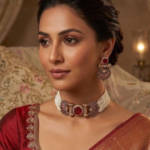 May include: A woman wearing a matching jewelry set. The set includes a white beaded choker necklace with a large red square center stone and matching earrings. The earrings and necklace have a gold-colored metal design.