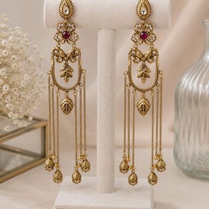 May include: A pair of long, antique gold-toned dangle earrings featuring a teardrop-shaped top, a central flower motif with a deep red stone, and a U-shaped frame with hanging chains and small, textured bell-shaped charms at the bottom.