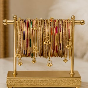 May include: A gold bangle stand displaying numerous bangles in various colours. The stand has a rectangular base with decorative detailing and a vertical frame holding the bangles. Some bangles have dangling charms.