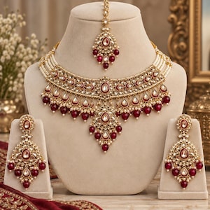 May include: A gold-toned jewellery set featuring a wide choker necklace, a pair of matching drop earrings, and a maang tikka. The pieces are adorned with teardrop-shaped red stones, clear crystals, and small white pearls in a layered design.