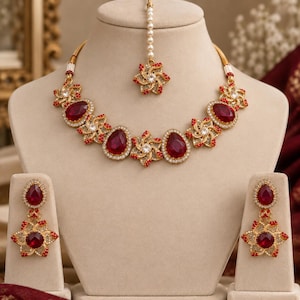 May include: A gold-toned jewelry set featuring a necklace, matching drop earrings, and a maang tikka. The pieces are adorned with large, oval-shaped deep red stones, smaller clear crystals, and tiny red beads arranged in star-like floral patterns.