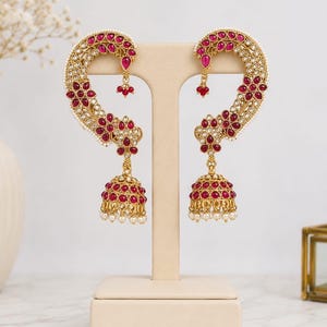 May include: Gold-coloured earrings with a curved design, set with red gemstones and small white pearls. The earrings have a bell-shaped drop with more red stones and pearls, displayed on a neutral stand.