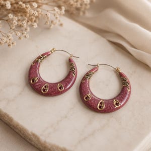 May include: A pair of crescent-shaped hoop earrings featuring a mauve marbled enamel finish with gold-toned metal accents. Each earring is decorated with three small, teardrop-shaped red gemstones set in gold frames and intricate metal detailing.