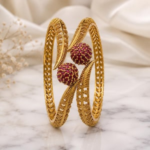Peut inclure: Un bracelet jonc en métal doré présentant une surface texturée complexe et deux accents sphériques proéminents incrustés de petites pierres roses éclatantes. Le design comprend des éléments incurvés en forme de feuille entourant les sphères.