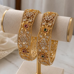 May include: Two gold-toned bangles featuring intricate filigree metalwork, clear sparkling stones, and floral motifs. One bangle is accented with red circular stones, while the other features green circular stones at the centre of each flower.
